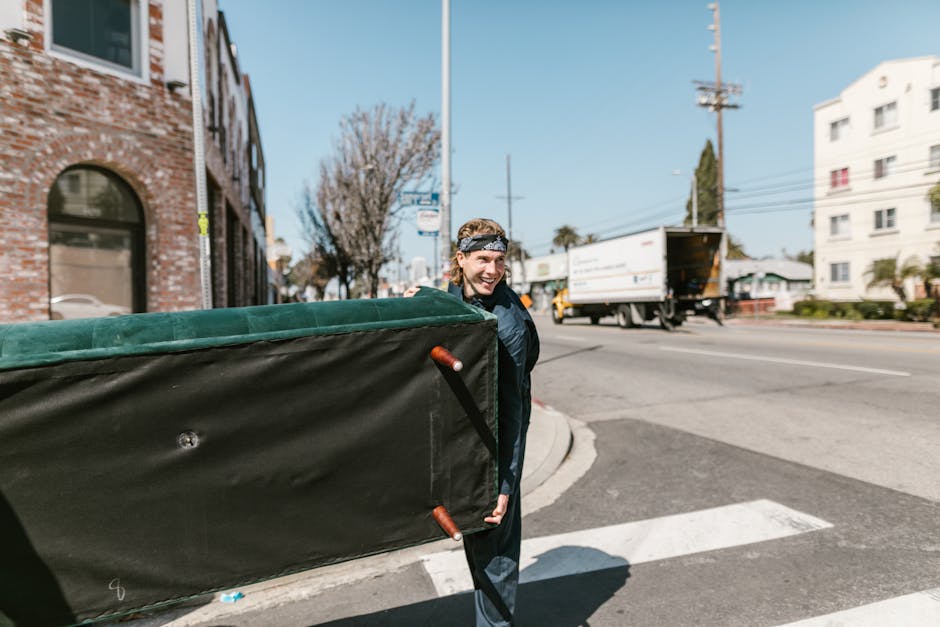 A man with light skin, wearing a black jacket and a grey patterned headband, is carrying a large, rectangular moving box wrapped in black plastic with orange handles, during a house removal process. He is standing on a city street near a corner, holding the box with both hands, and smiling. The background features a brick building with arched windows, a leafless tree, a white multi-storey building, and a delivery truck driving past in the distance. The scene is bathed in daylight with clear blue skies, indicating daytime, and suggests an outdoor loading or packing stage of a home relocation. Man with Van Haringey, a professional removals company, is involved in furniture transport and packing and moving services, assisting with the logistical aspects of house removals, including loading large boxes onto a van. The environment shows typical urban elements like traffic signals, road markings, and pavement areas.