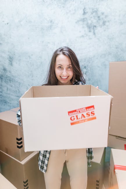 A woman with long brown hair and a friendly expression, standing indoors against a textured light blue wall, holding a large cardboard box with a red and white sticker that reads 'GLASS' in capital letters. She is dressed casually in a plaid shirt and light-colored trousers. Surrounding her are several other cardboard boxes, some sealed with tape and others open, prepared for a home relocation. The scene conveys the packing process involved in house removals, with the woman likely representing a professional moving service. The setting suggests an indoor area used for sorting and packing household items, emphasizing careful handling of fragile objects during furniture transport and moving logistics as part of a comprehensive removals service, as offered by Man with Van Haringey.