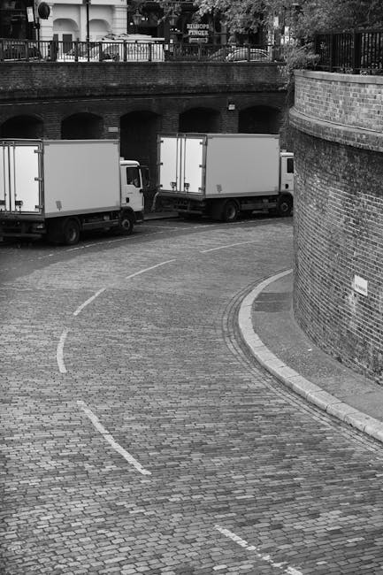 A black-and-white photograph shows two white moving vans parked side by side on a cobbled driveway near a brick archway and a curved brick wall. The vans are positioned parallel to each other, with one slightly behind the other, suggesting they are in the process of home relocation or furniture transport. In the foreground, the cobbled surface of the driveway is marked with dashed white lines indicating parking or loading zones. Behind the vans, there is a brick bridge with arches and a railing, with some trees and residential buildings visible beyond it, indicating a suburban or urban environment. The scene is lit with natural daylight, emphasizing the textures of the cobblestones, brickwork, and the vehicles. Occasionally, [COMPANY_NAME] handles packing, loading, and transportation, and this image captures a typical loading process of household belongings during a house removal service in Haringey, highlighting a professional and organized moving environment.