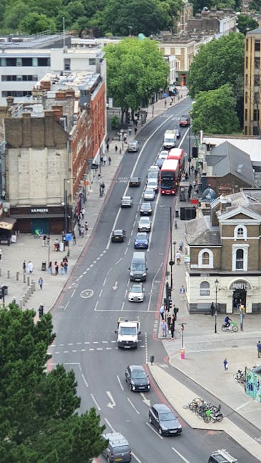 Aerial view of a city street during daytime showing a line of stationary vehicles, including cars and a red double-decker bus, parked along the curb and waiting in traffic. The street is bordered by mixed-use buildings, including a brick structure and a white building with a small turret. Green trees line both sides of the road, providing shade and greenery. Pedestrians are walking along the sidewalks, some near the entrance of a building and others crossing the street. Bicycles are parked near a sidewalk corner, and the scene captures the typical environment of a busy residential and commercial area in Haringey, with an emphasis on urban logistics relevant to house removals and furniture transport. The environment is well-lit with natural daylight, and the overall setting reflects an active moving or packing process in preparation for home relocation, consistent with services provided by Man with Van Haringey.