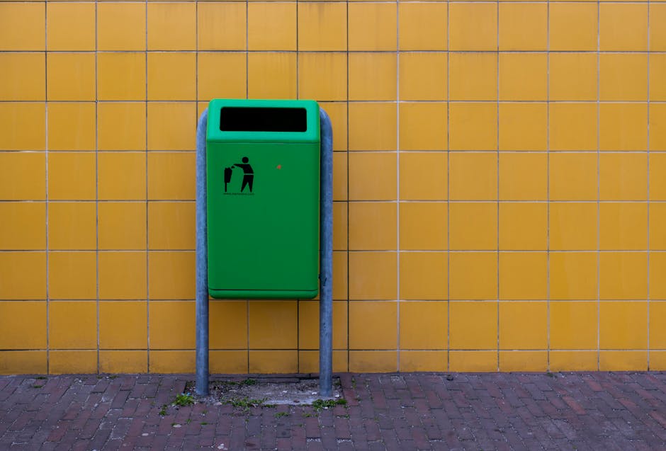 A green waste bin with a black pictogram indicating and a small red light, mounted on two vertical grey metal posts, positioned against a yellow tiled wall outdoors. The bin is designed for rubbish di
