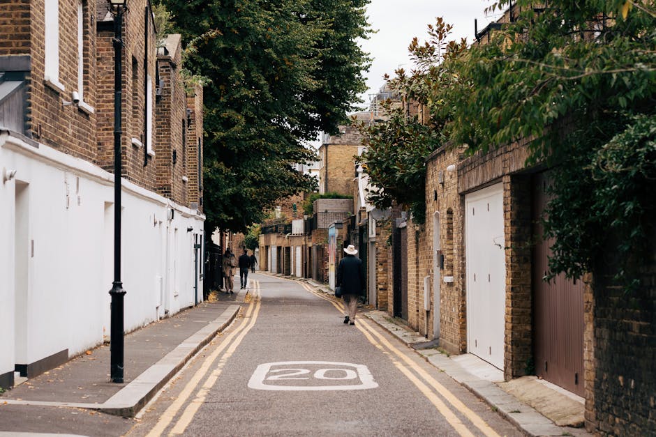 A narrow residential street in Crouch End, Haringey, with brick houses on both sides and a white-painted garage door on the right. The street features a 20 mph speed limit marked on the asphalt, with double yellow lines along the curbs. A man wearing a dark coat and a white hat is walking along the pavement, while further down, two people are seen moving away. The scene includes trees with foliage extending over the street, and the lighting suggests an overcast day, typical of an urban house removal or relocation setting. The environment appears calm and typical for a household furniture transport process or packing and moving activities in a residential area, with no visible moving equipment or furniture in the current scene, but the context indicates preparation for or completion of a home relocation.