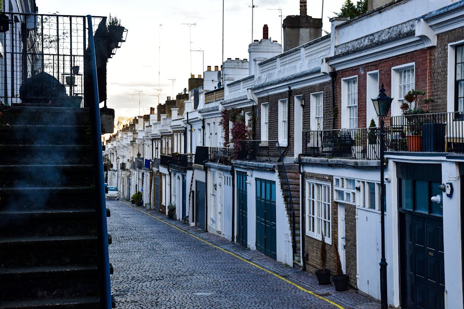 A narrow residential street in Crouch End with a cobblestone pavement, lined by terraced brick houses featuring white and dark-colored doors and window frames. Some houses have small front gardens with potted plants and wrought iron railings, while others have external staircases leading to upper floors. The sky is overcast, and the soft evening light casts subtle shadows. On the left side, a black metal staircase with steps and railing is visible, possibly leading to an upper apartment. The street appears quiet and clean, with a few small cars parked along the curb. This setting reflects an urban neighbourhood typical for house removals and home relocation services in Haringey, where careful furniture transport and packing are necessary for moving house. Man with Van Haringey, specialising in removals, operates within this environment, ensuring efficient moving logistics in narrow streets like this.