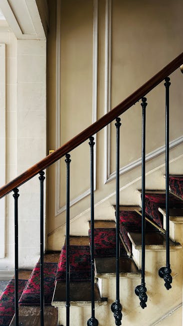 Inside a residential property, a staircase with narrow Victorian-style steps, covered partially by a red patterned carpet, leads upward. The staircase features a dark wooden handrail supported by black metal balusters. The walls adjacent to the stairs are paneled with cream-colored wainscoting, and the surrounding environment is softly lit, highlighting the texture of the wood and the fabric on the steps. Part of the staircase is visible through an open doorway on the left, with a glimpse of the hallway beyond. This scene is representative of home relocation or furniture transport processes often involved in house removals, such as those offered by Man with Van Haringey, especially when navigating challenging narrow staircases typical of Victorian houses.