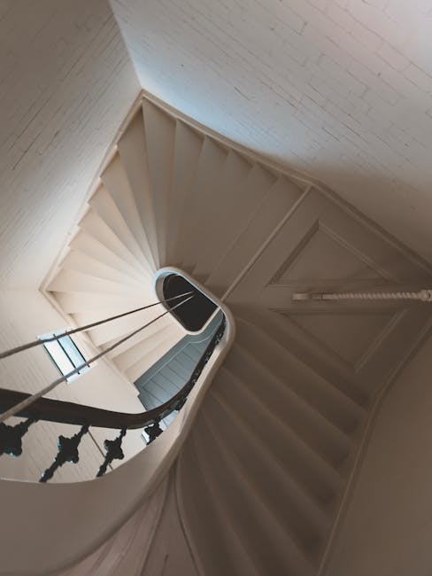 A photograph capturing a narrowly winding staircase inside a residential property, with white painted wooden stairs and a curved black metal balustrade featuring decorative spindles. The staircase ascends from the bottom left corner to the top, with a small landing visible at the top. The surrounding walls are painted in a light neutral colour, and a window is partially visible at the bottom of the staircase, allowing natural light to illuminate the area. The ceiling at the top of the staircase is slightly angled. The image was taken during a home relocation process, with the staircase likely being part of a house being prepared for furniture transport or packing and moving services, as offered by Man with Van Haringey.