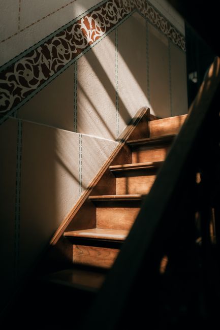 Inside a residential property, a staircase with narrow Victorian-style steps, covered partially by a red patterned carpet, leads upward. The staircase features a dark wooden handrail supported by black metal balusters. The walls adjacent to the stairs are paneled with cream-colored wainscoting, and the surrounding environment is softly lit, highlighting the texture of the wood and the fabric on the steps. Part of the staircase is visible through an open doorway on the left, with a glimpse of the hallway beyond. This scene is representative of home relocation or furniture transport processes often involved in house removals, such as those offered by Man with Van Haringey, especially when navigating challenging narrow staircases typical of Victorian houses.