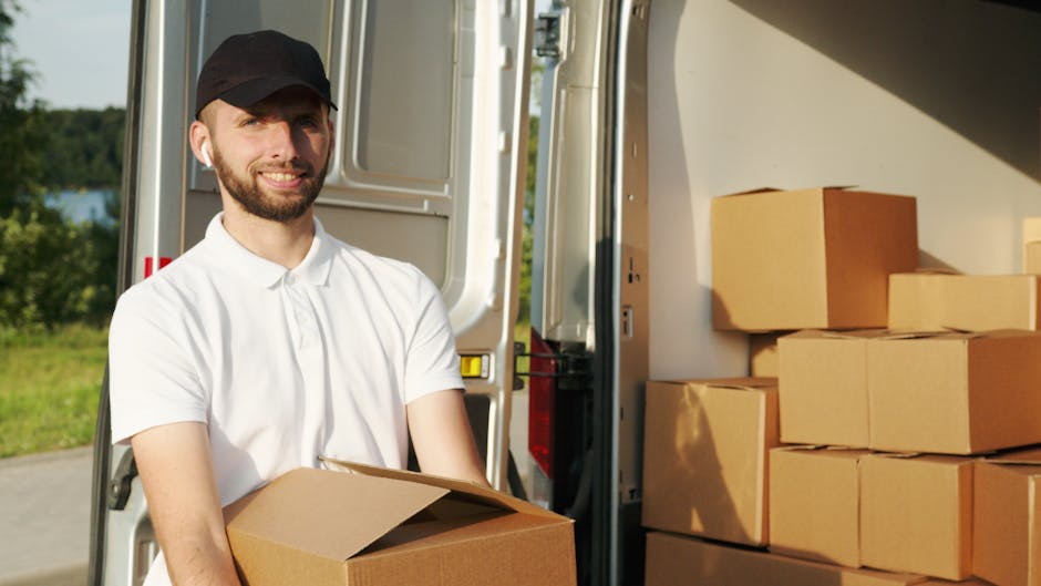 A man with a beard, wearing a white polo shirt and a black cap, is standing outside near an open moving van during daylight. He is holding a cardboard box and smiling at the camera. Inside the van, several neatly stacked cardboard boxes are visible, along with a few blankets or protective wrapping materials on the floor. The open van door reveals a ramp or loading area, with some greenery and trees in the background indicating an outdoor setting. The scene depicts the process of packing or unloading boxes as part of a home relocation or furniture transport service, supported by the presence of the moving vehicle and packing materials. Man with Van Haringey is performing a loading or unloading task as part of professional removals, emphasizing the reliable handling of packing and moving logistics.