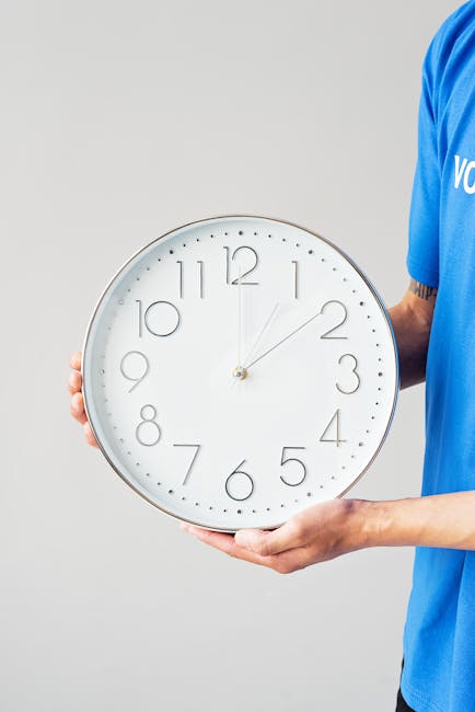 A person wearing a blue shirt is holding a large white wall clock with a silver frame. The clock has minimalistic black numerals and slim black hour and minute hands, showing the time as approximately 11:03. The person is gripping the left side of the clock with their left hand and supporting the bottom with their right hand, standing against a neutral grey background. The scene appears to be indoors, possibly in a home or office environment, and the clock is positioned to demonstrate time display, which is relevant to house removals and timing logistics. This image emphasizes the importance of time management during home relocation processes, handled by companies such as Man with Van Haringey, which provides removals services.