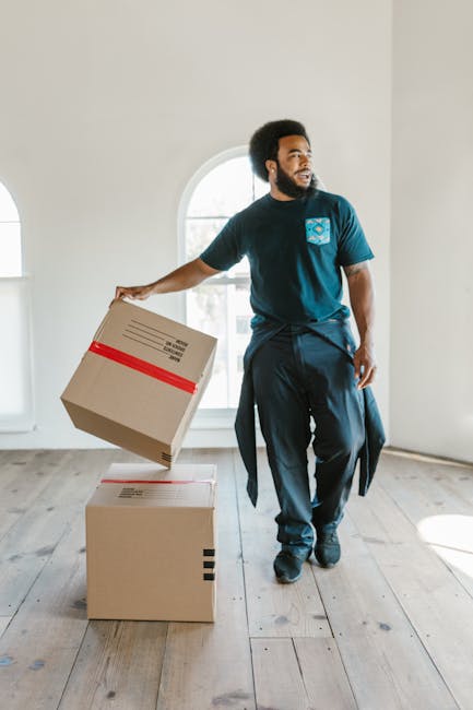 A man with a dark curly afro hairstyle and beard, dressed in a blue T-shirt and black trousers, is inside a bright, spacious room during a furniture transport process. He is holding a cardboard box wrapped with red tape in his right hand, preparing to place it on top of another box on the light wooden floor. Behind him, a large arched window allows natural light to illuminate the space, which has white walls and minimal furnishings, reflecting an ongoing home relocation. The scene suggests a professional moving service, with a focus on packing, handling, and furniture transport as part of house removals. The environment and equipment, including the cardboard boxes, indicate careful packing and moving logistics by [COMPANY_NAME], supporting efficient packing and packing and moving processes involved in house removals or relocation services.