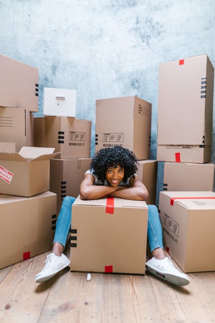 A young woman with curly black hair, dressed in casual clothing including a white top, blue jeans, and white sneakers, sits on a light wooden floor inside a room filled with numerous cardboard boxes of various sizes, some with red and black tape securing the closures. The boxes are stacked against light blue textured walls, creating a cluttered environment typical of a home relocation or moving process. The woman is smiling and leaning forward with her arms resting on a large box at her front, suggesting a break during packing or unpacking activities. The scene includes a mix of open and closed boxes, indicating packing and organizing phases, and the natural lighting illuminates the space evenly. This image, associated with house removals and furniture transport services offered by Man with Van Haringey, captures a moment of packing or moving logistics essential to home relocations.