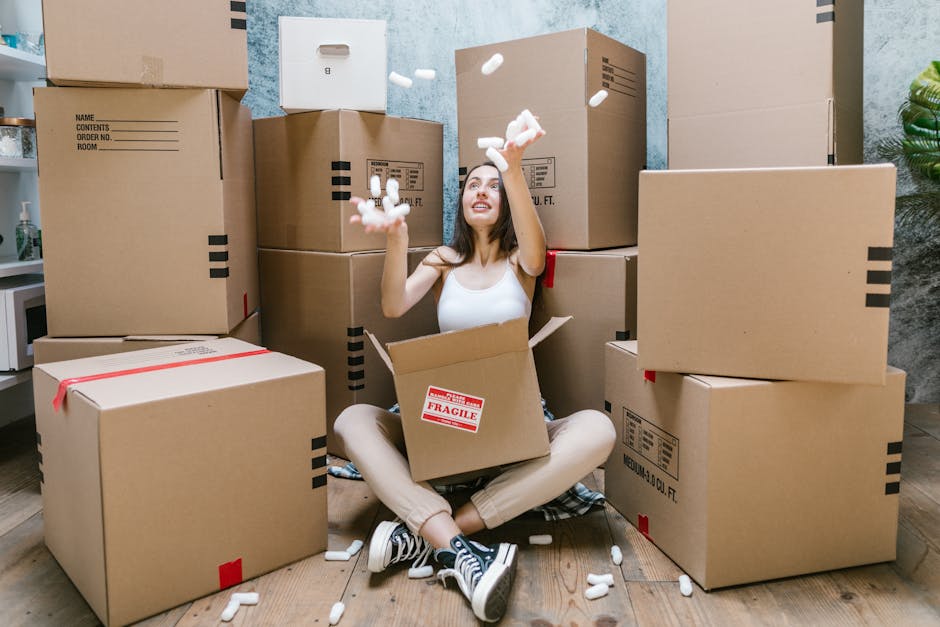 A young woman with long dark hair, dressed in a white sleeveless top and beige trousers, is sitting cross-legged on a wooden floor inside a room, surrounded by large cardboard boxes and packing materials. She is smiling and joyfully throwing or catching white packing peanuts above her head into the air. The boxes are various sizes, some sealed with red tape, and have labels indicating contents or fragile items. Behind her, the room features a textured grey wall, a small white cabinet on the left with a plant and other items, and a large green plant on the right. The scene suggests a home relocation process involving packing and moving furniture, with a focus on careful handling and transportation of household items. The environment is well-lit with natural or artificial light highlighting the packing activity, and the setting reflects the interior of a house being prepared for a removal service, such as those offered by Man with Van Haringey.