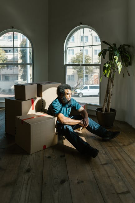 A young woman with curly black hair, dressed in casual clothing including a white top, blue jeans, and white sneakers, sits on a light wooden floor inside a room filled with numerous cardboard boxes of various sizes, some with red and black tape securing the closures. The boxes are stacked against light blue textured walls, creating a cluttered environment typical of a home relocation or moving process. The woman is smiling and leaning forward with her arms resting on a large box at her front, suggesting a break during packing or unpacking activities. The scene includes a mix of open and closed boxes, indicating packing and organizing phases, and the natural lighting illuminates the space evenly. This image, associated with house removals and furniture transport services offered by Man with Van Haringey, captures a moment of packing or moving logistics essential to home relocations.